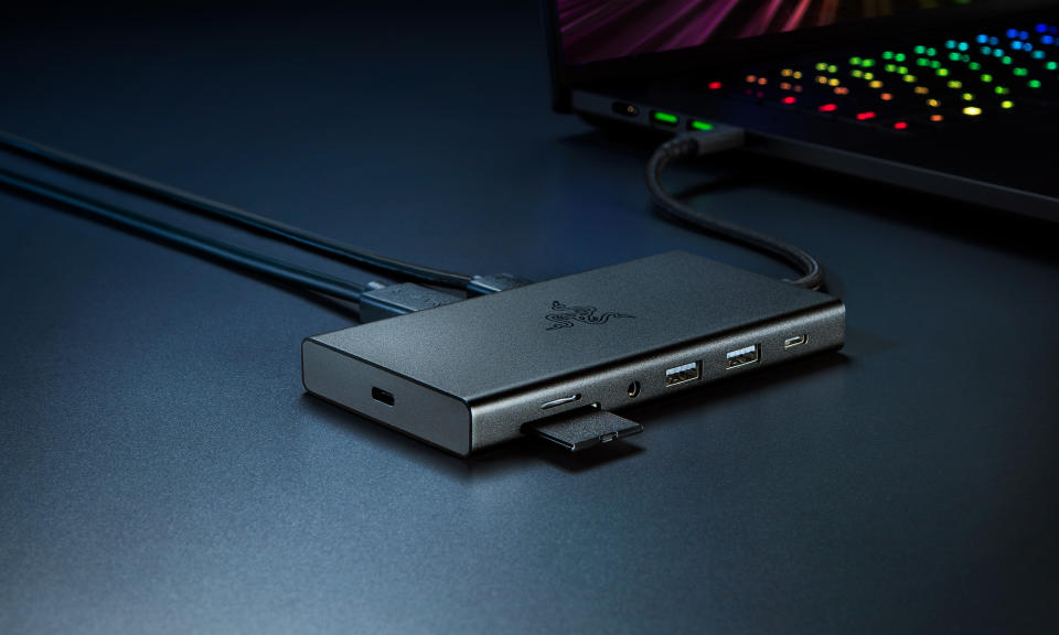 Productmarketingafbeelding voor de Razer USB C Dock.  Het centrum bevindt zich op een bureau waarachter een gaming-laptop staat.  Het heeft veel open poorten en een half-ready SD-kaart.  Het staat op een donkerblauw bureau met dramatische tinten.