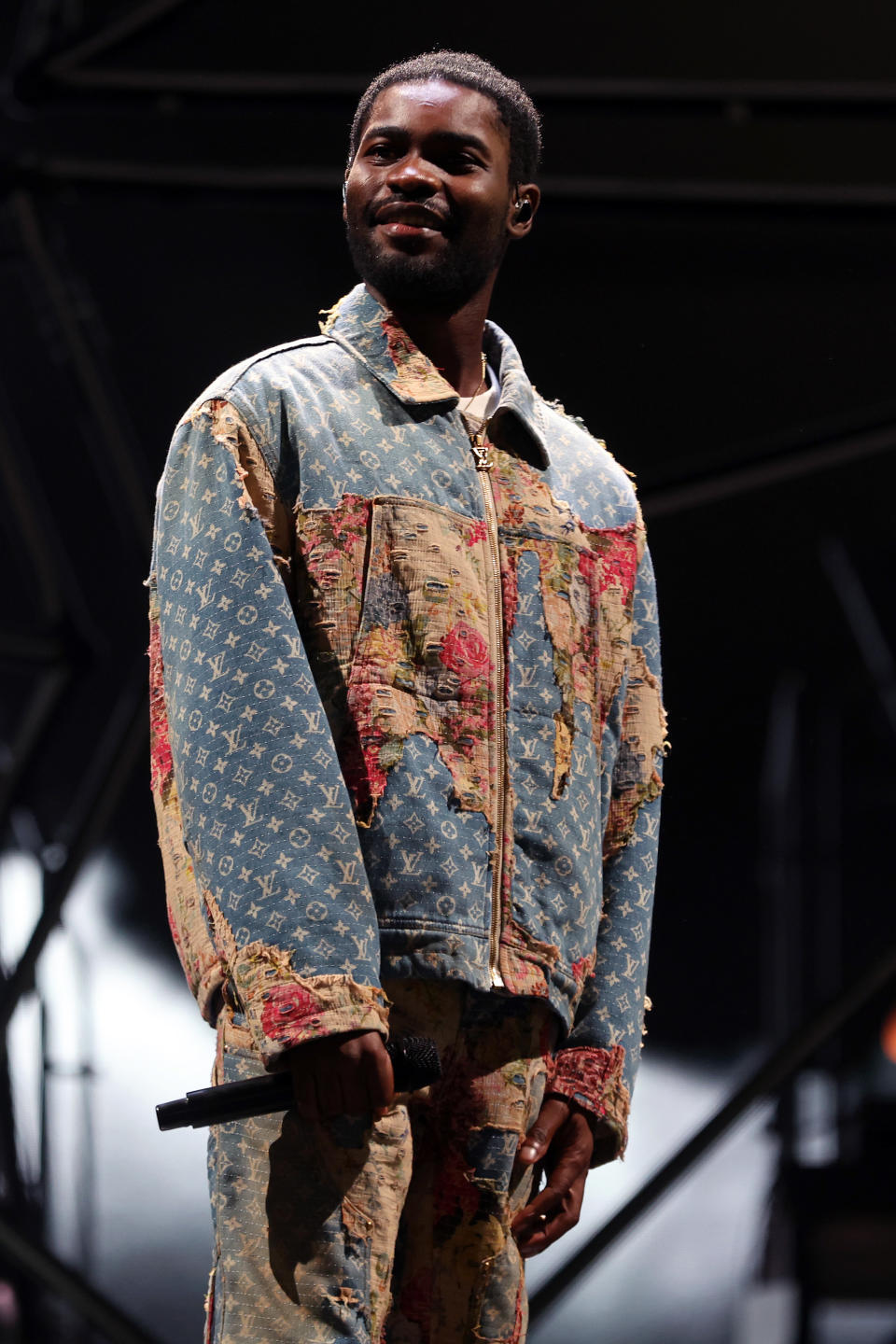 Dave stands on stage holding a microphone during a performance. Dave is wearing a denim Louis Vuitton logomania branding