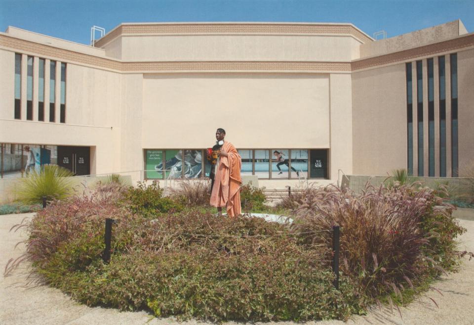 A man stands in front of a building holding a cup and a blanket.