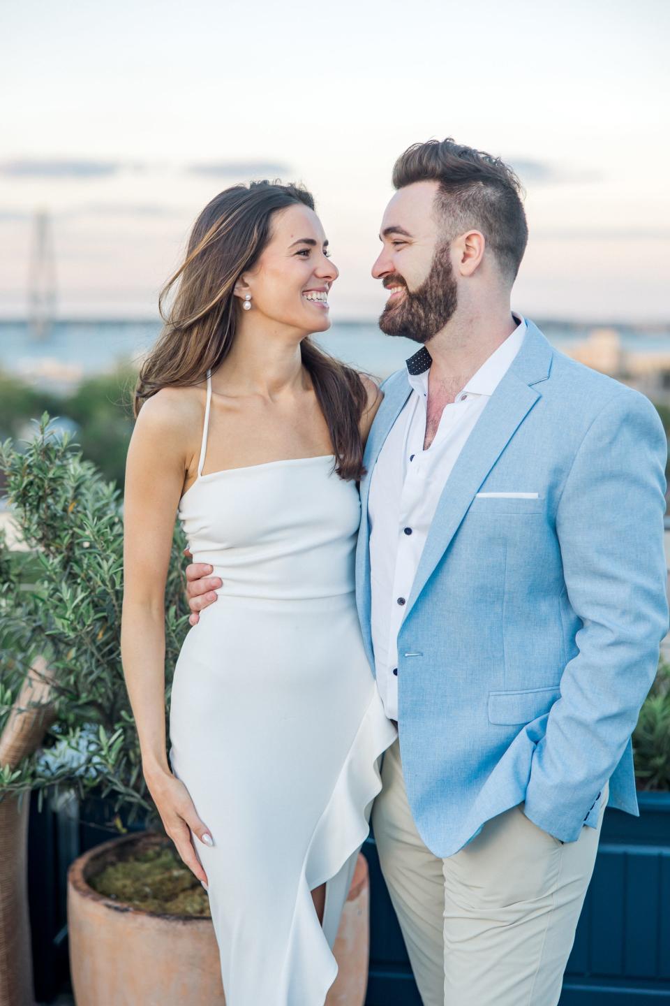 A bride and groom smile at each other in front of a sunset.