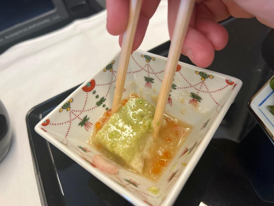 The author using chopsticks to pick up green pea tofu from a square, white dish.