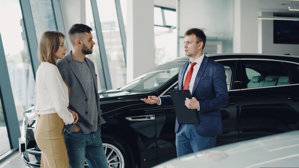 Interested young couple man and woman are talking to manager in car dealership while he is telling them about luxury auto model. Beautiful people, transportation and business concept.