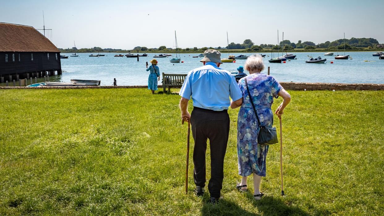  An elderly man and woman walk arm in arm and with canes towards a harbor full of sailboats. 
