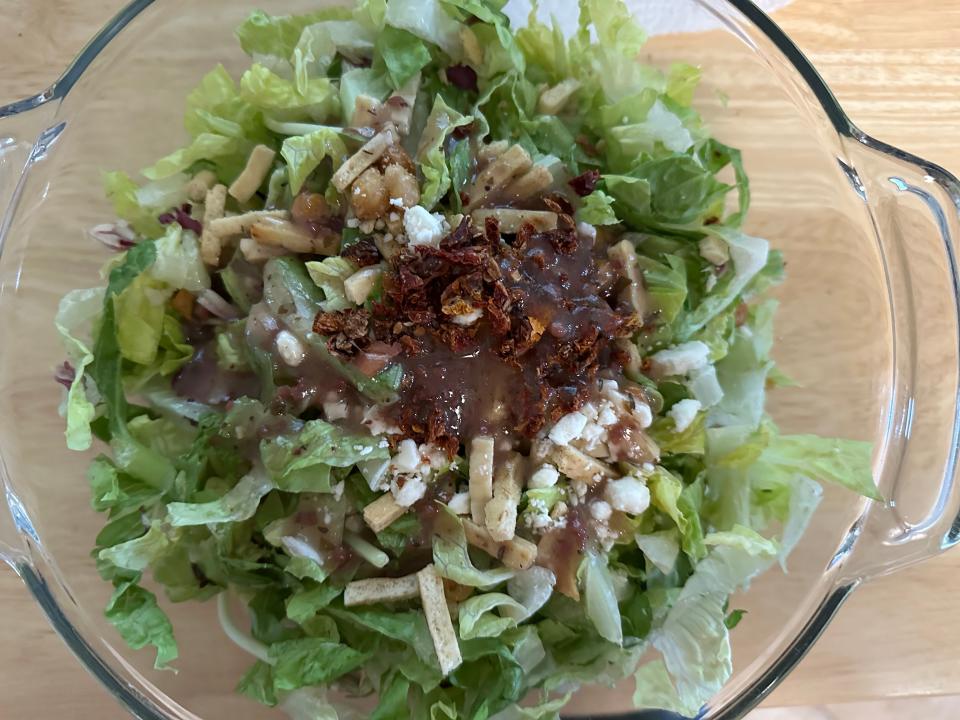 green salad topped with dried tomato pieces, small flatbread sticks, and feta crumbles in glass bowl