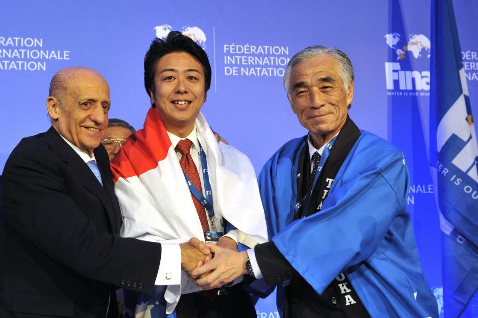 FILE - In this Jan. 31, 2016 file photo, from left, President of International Swimming Federation, FINA, Julio Maglione, President of the Japanese Swimming Federation Tsuyoshi Aoki and Mayor of Fukuoka, Japan, Soichiro Takashima shake hands after Fukuoka was announced to host the 2021 FINA world aquatics championships during a meeting of FINA leaders in Budapest, Hungary. On Monday, March 30, 2020, the Tokyo Games were pushed back a full year by the coronavirus pandemic, to the same period of the biannual aquatics, causing the swimming’s governing body to go back to the drawing board to figure out when to hold its next world championships. (Attila Kovacs/MTI via AP, file)