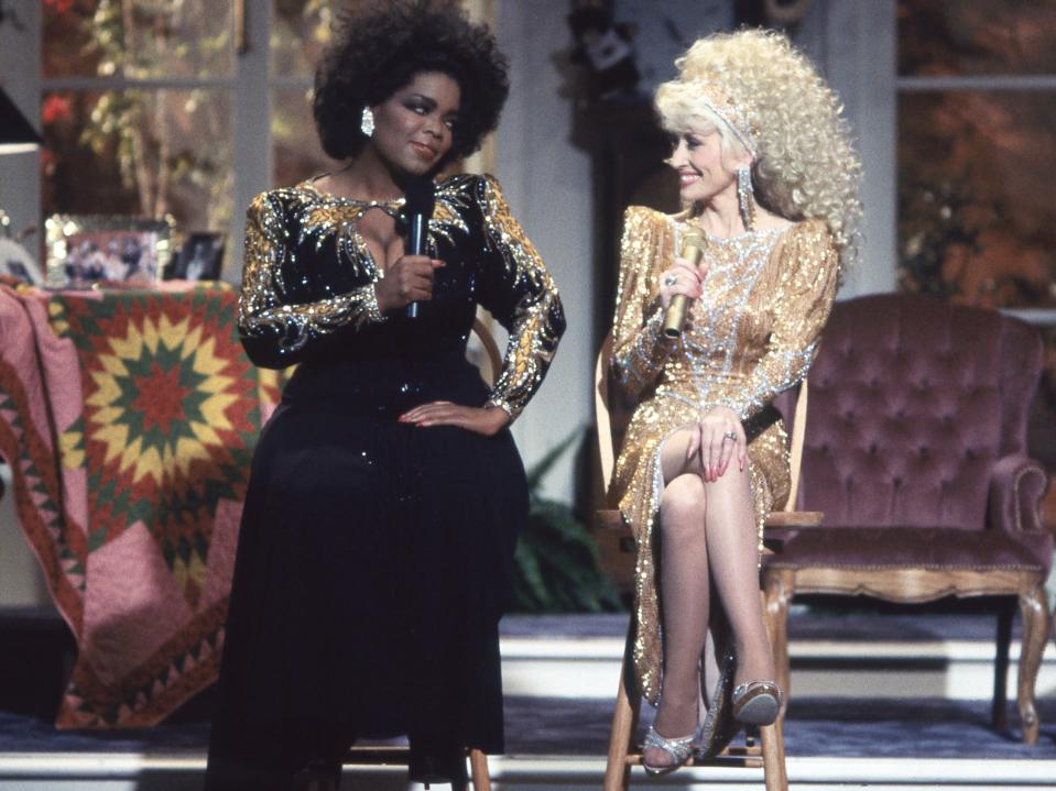 Oprah Winfrey in a black gown with gold detailing and Dolly Parton in a gold dress sitting on stools next to each other.