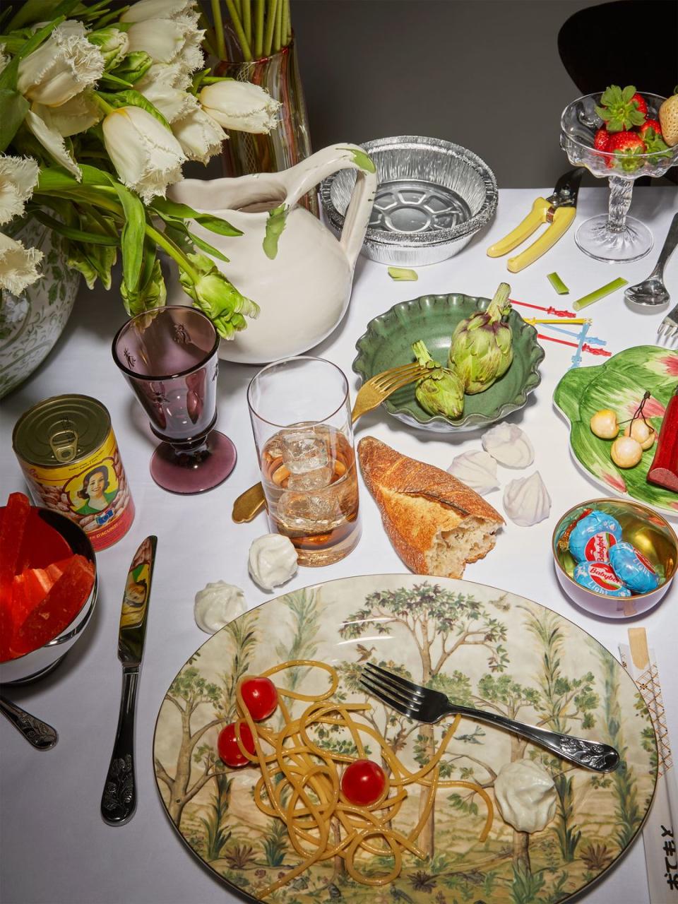 a table is filled with food and plates of all colors and sizes and some silverware and glassware and a white pitcher with faded white tulips