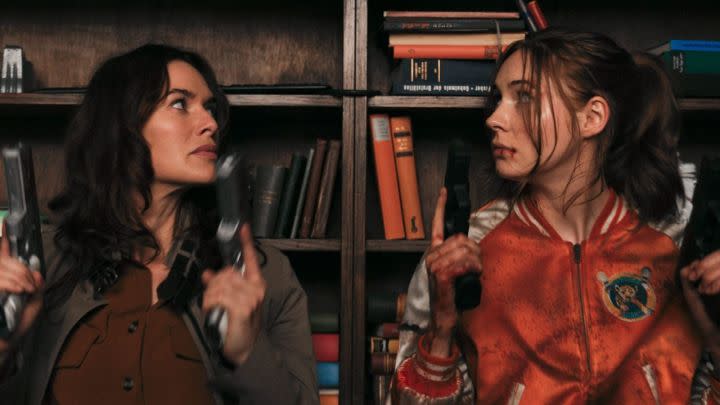 Lena Headey and Karen Gillan as Scarlet and Sam holding guns and taking cover behind a bookshelf in the film Gunpowder Milkshake.