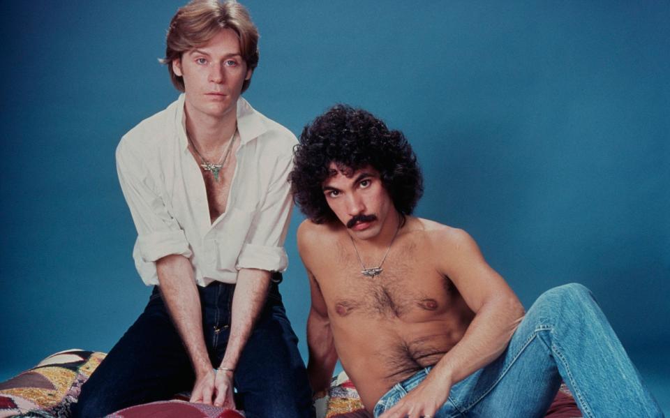 American rock and soul duo Hall & Oates (American singer-songwriter and musician Daryl Hall, wearing a white shirt, open at the collar, and American singer-songwriter and guitarist John Oates who poses shirtless) in a studio portrait, against a blue background - Lynn Goldsmith/Getty Images