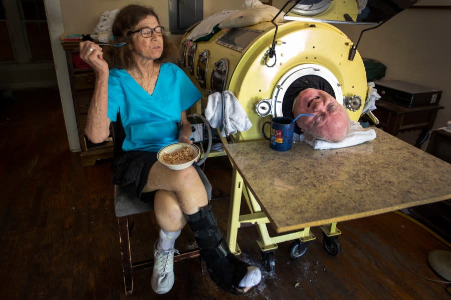 FILE – In this Friday, April 27, 2018 photo, attorney Paul Alexander chats with caregiver and friend Kathryn Gaines as he drinks coffee and she eats breakfast beside his iron lung at his home in Dallas. Alexander died Monday, March 11, 2024 at a Dallas hospital, said Daniel Spinks, a longtime friend. He said Alexander had recently been hospitalized after being diagnosed with COVID-19 but did not know the cause of death. (Smiley N. Pool/The Dallas Morning News via AP)