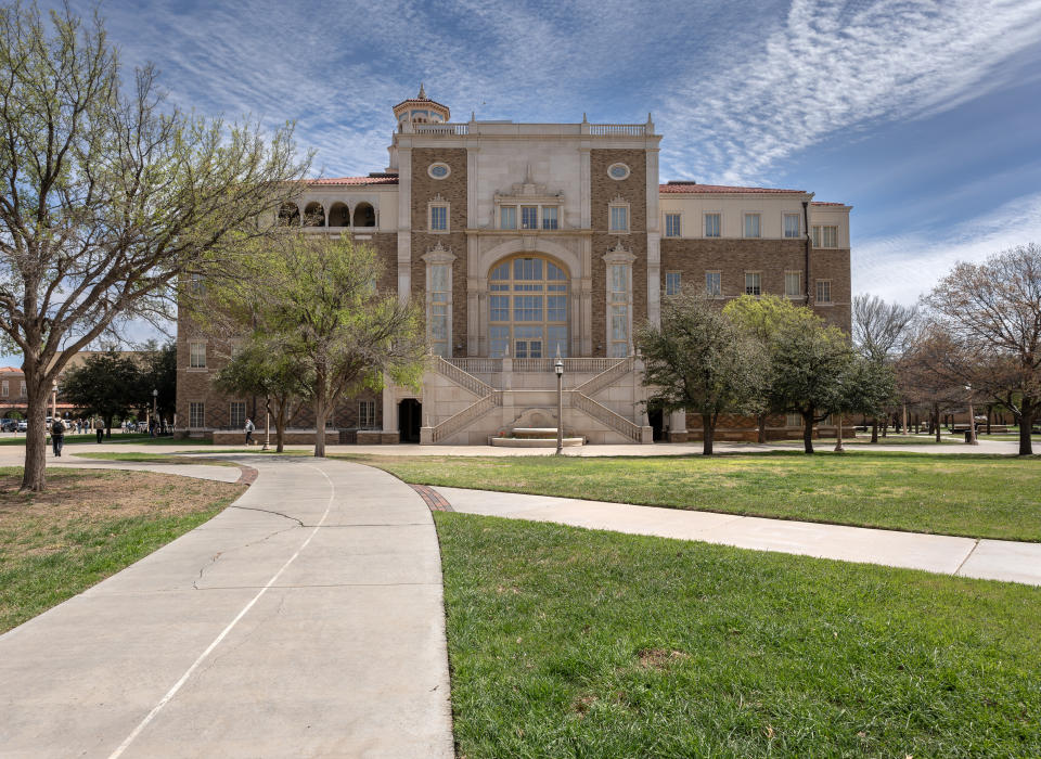 A large, rectangular building with a grand entrance, surrounded by trees and a large open lawn. A paved pathway leads up to the building