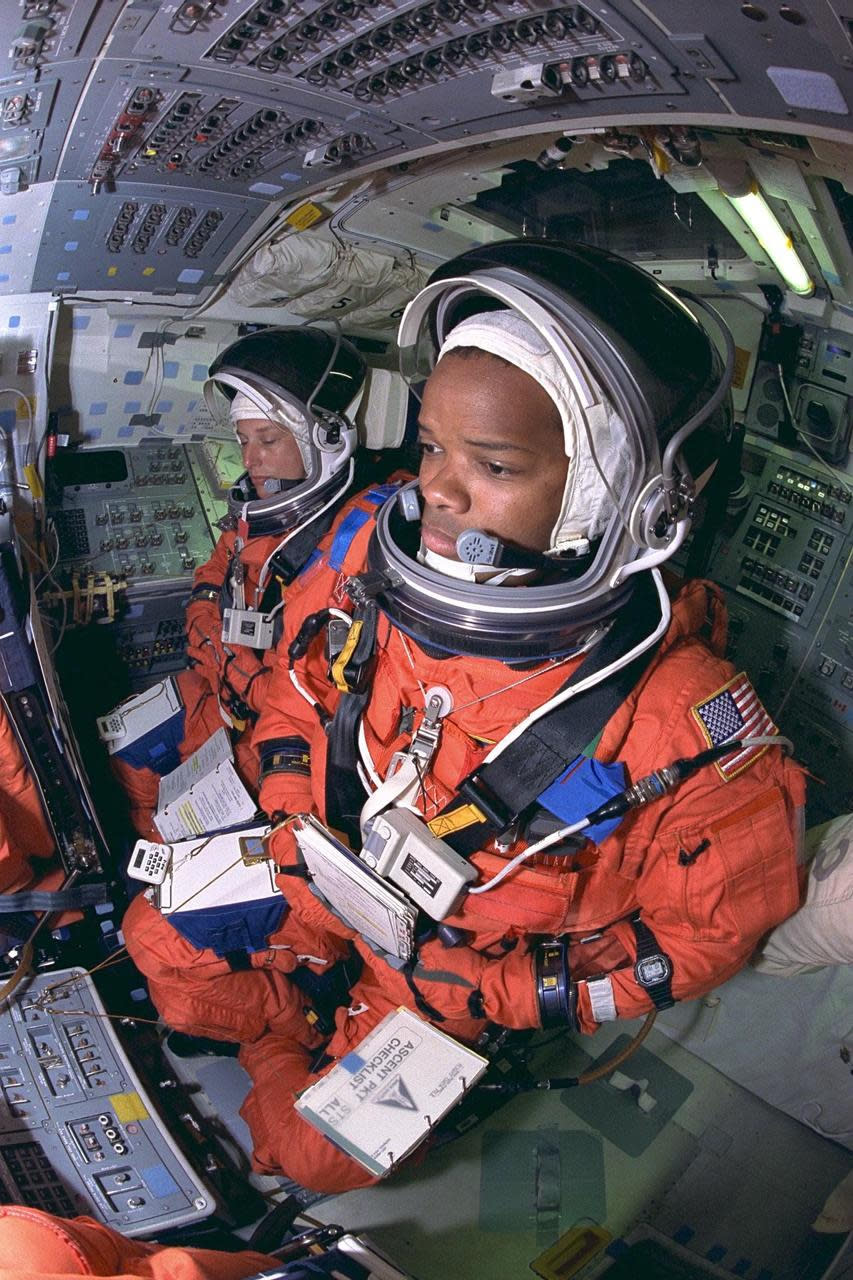 two astronauts in orange sit at a cockpit full of switches and buttons.