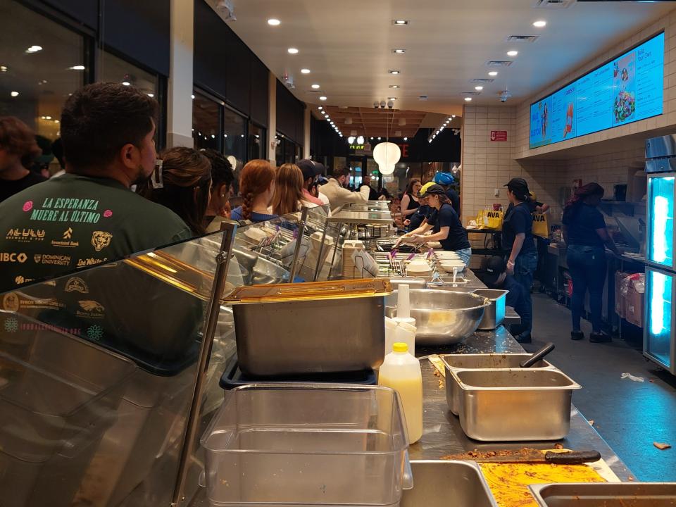 The interior of the Cava restaurant in Chicago, showing the counter which is full of ingredients as well as a line of customers waiting to order