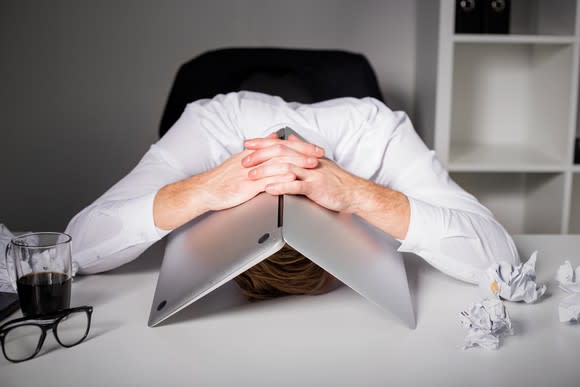 Man with head on desk and laptop over his head.