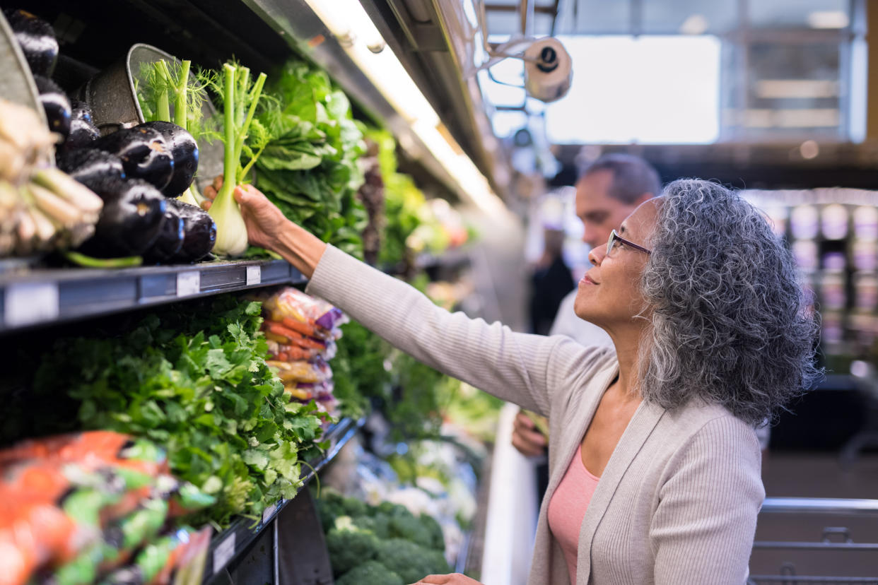 A senior couple pick out fresh produce together at the supermarket. The focus is on her as she reaches up to inspect veggies.