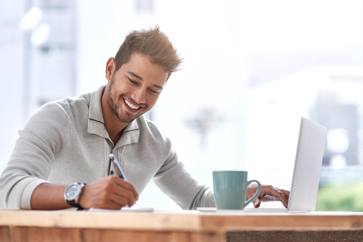 Smiling young man writing with a pencil and other hand is on a laptop on a wooden desk, a seafoam coffee cup, with a large window in the office blurred in the background