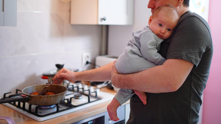 A person cooks on a gas stove while holding a baby.