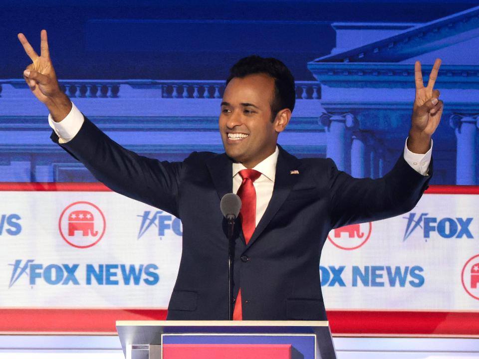 Vivek Ramaswamy grins and flashes the V sign with both hands on a debate stage.