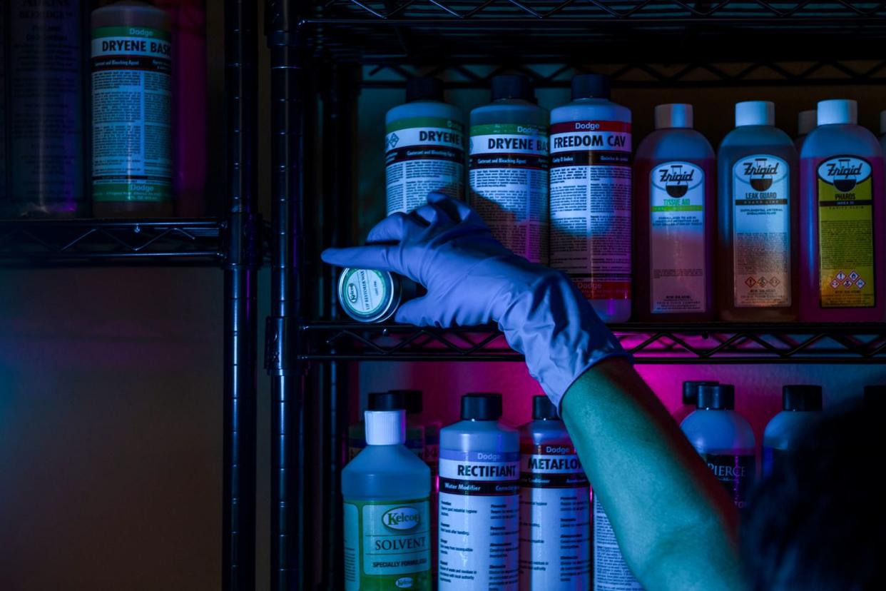 gloved hand reaching for tin of lip restorer wax on shelf of embalming fluids