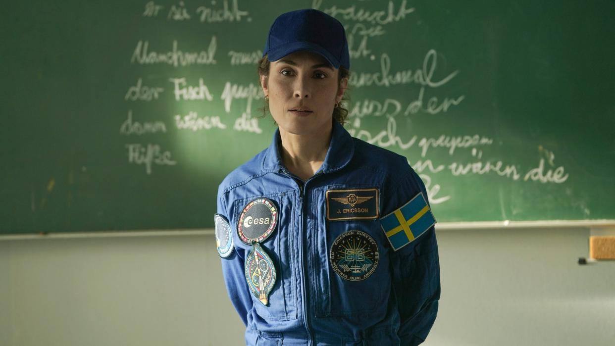  A woman wearing a blue astronaut jumpsuit decora ted with several different patchers, including a Swedish flag. She is also wearing a blue baseball cap. She is standing in front of a black board with some chalk writing on. 