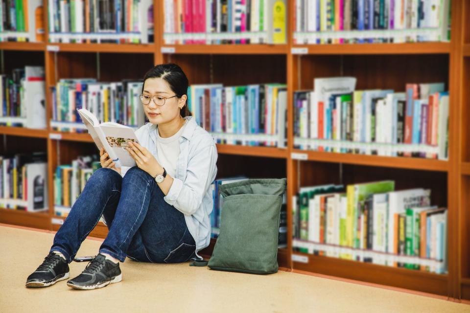 asian female adult student sitting on floor reading a book in a library