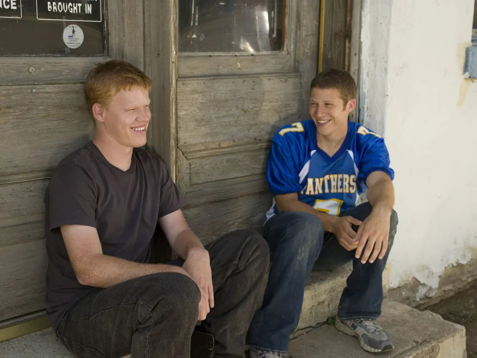 jesse plemons and zach gilford in character for friday night lights, they sit on a door step and smile while looking away from the camera, plemons wears a dark outfit and gilford wears a blue jersey and jeans