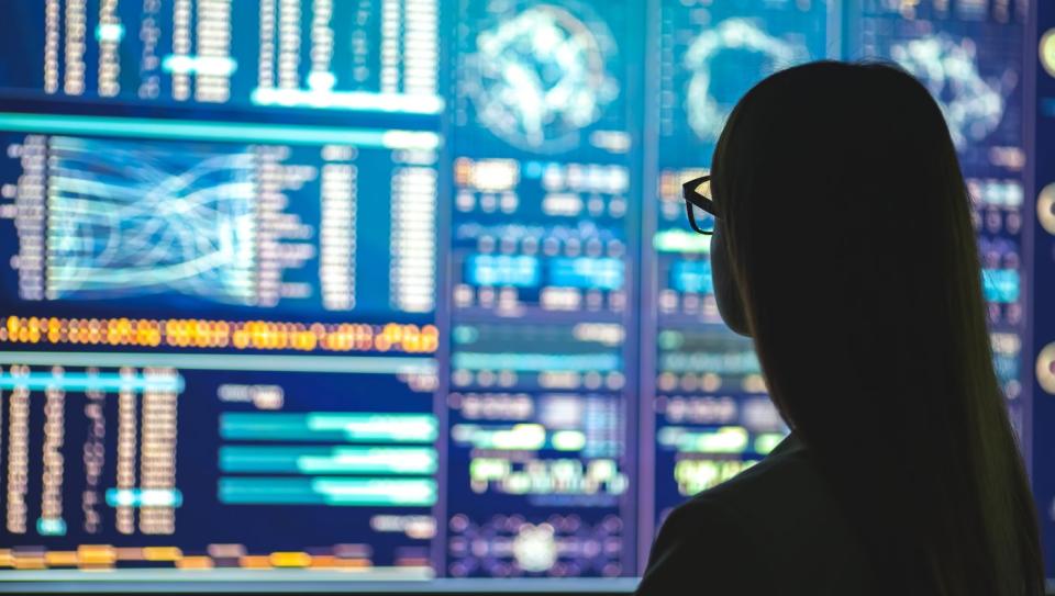 A businessman standing near several computers displaying various graphs.