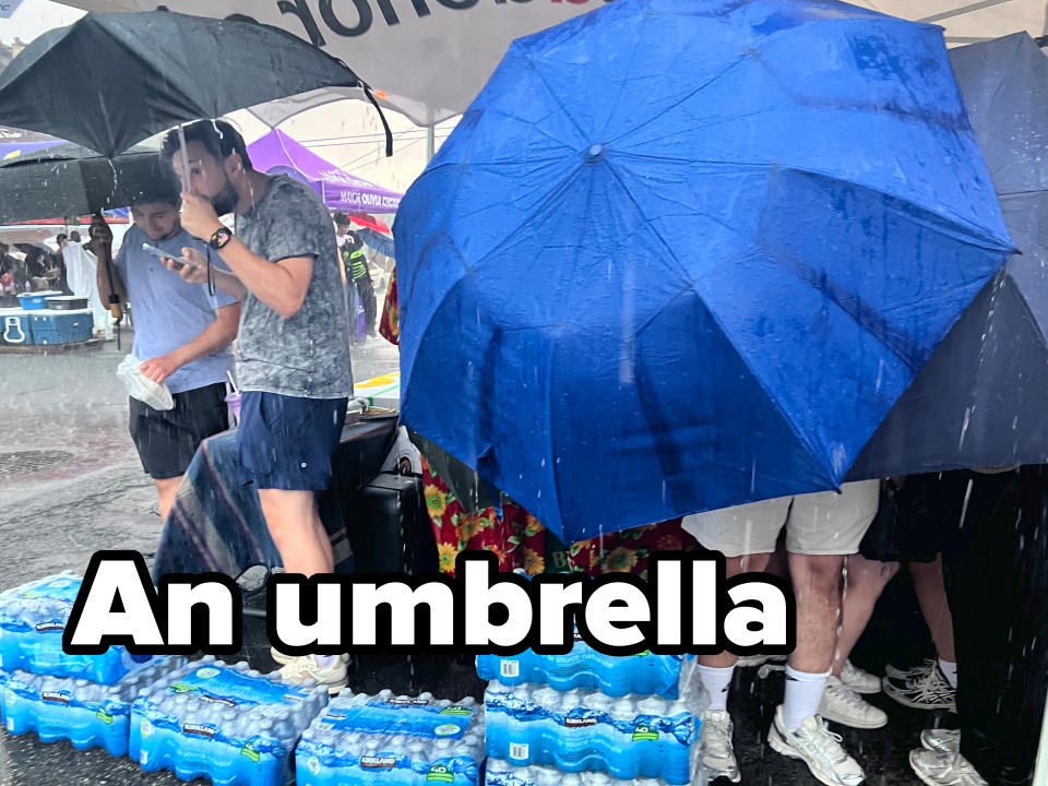 People standing under umbrellas in heavy rain, with multiple packs of bottled water at their feet