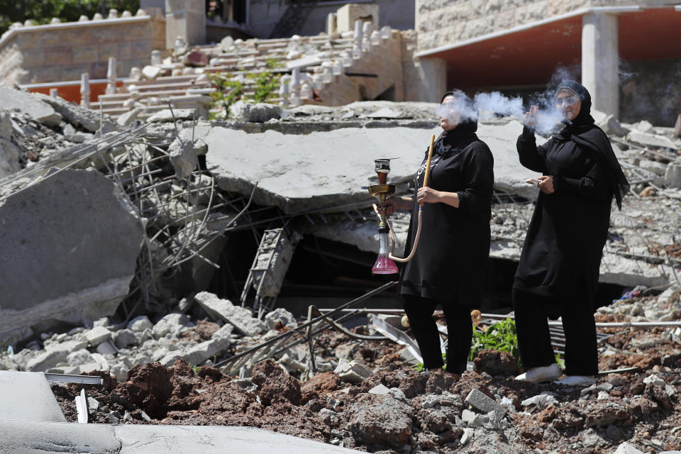 A Lebanese woman smokes a water pipe as another woman flashes a victory sign, while standing on the rubble of a destroyed house that was hit by an Israeli airstrike, in Aita al-Shaab, a Lebanese border village with Israel, south Lebanon, June 29, 2024. (AP Photo/Mohammed Zaatari)