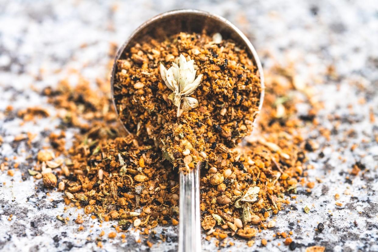 An antique spoon full of dukkah, a spice mix used in middle eastern cooking, spills out onto a stone table top