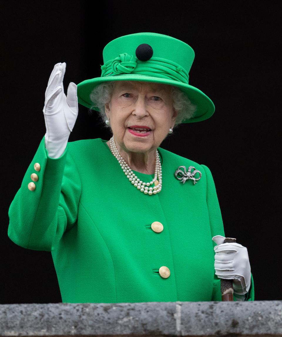 The Queen smiling and waving in a bright green coat with gold buttons and a matching green wide-brimmed hat. She's wearing perl earrings and necklaces, white gloves, and a diamond bow brooch.