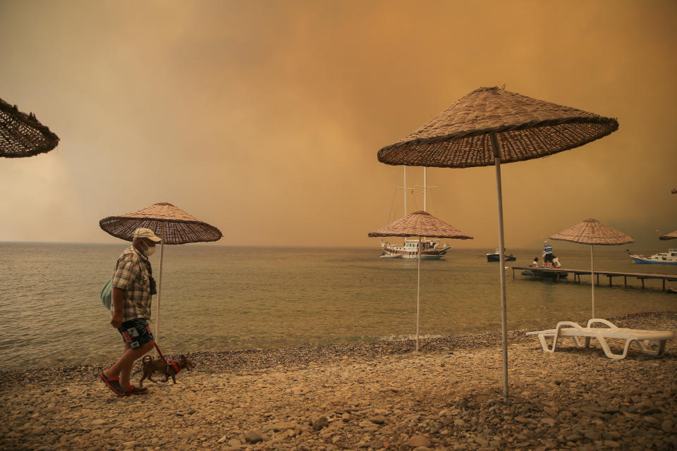 A man walks with his dog on the beach of smoke-engulfed Mazi area as wildfires rolled down the hill toward the seashore, in Bodrum, Mugla, Turkey, Sunday, Aug. 1, 2021. More than 100 wildfires have been brought under control in Turkey, according to officials. The forestry minister tweeted that five fires are continuing in the tourist destinations of Antalya and Mugla. (AP Photo/Emre Tazegul)