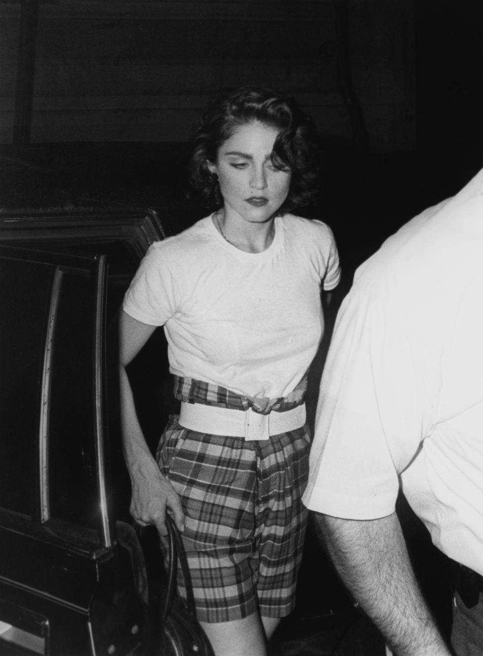 Madonna wearing a white t-shirt and plaid shorts, stepping out of a car. A person in the foreground partially obscures the view