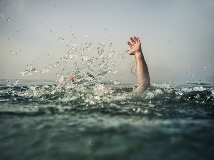 A person's hand reaching out of choppy water, indicating a potential drowning situation. The surrounding water is splashing