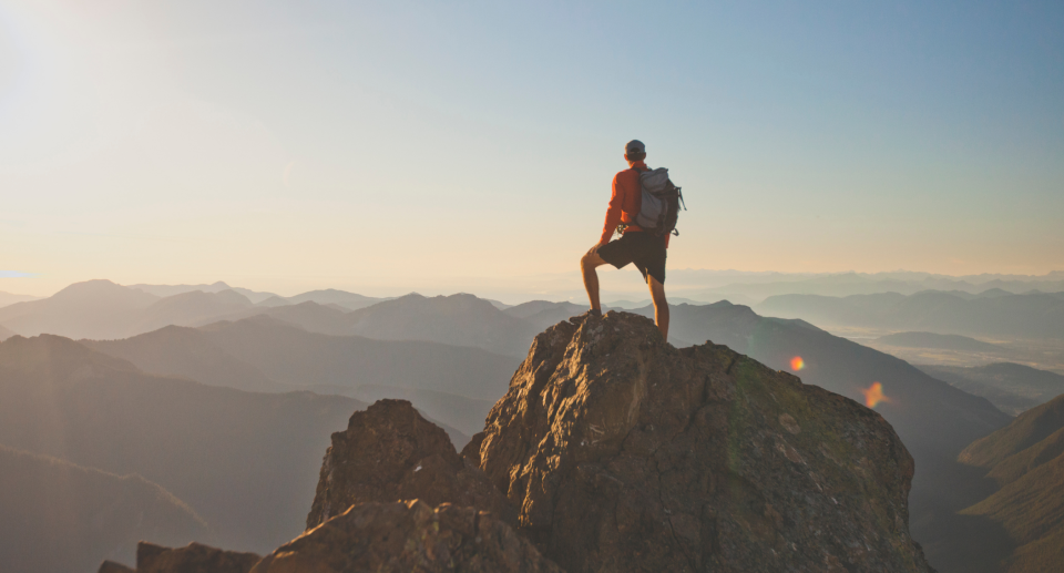 man in backpack standing on top of mountain over looking skyline, man hiking in mountains, Are you planning on hiking this summer? Here's what you'll need to keep in mind (Photo via Getty)