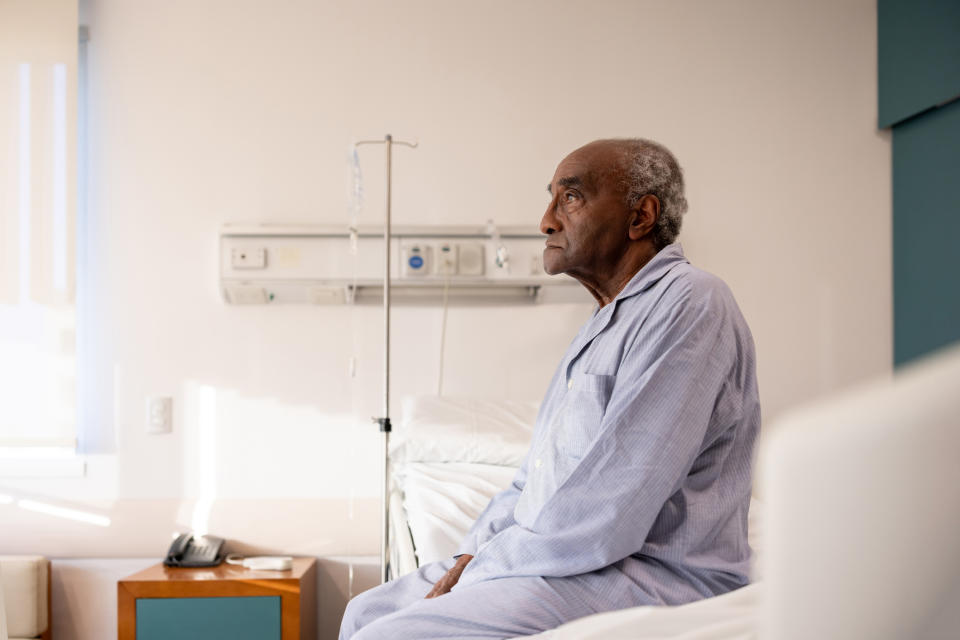 Elderly man in hospital gown seated by a bed, looking thoughtful, in a hospital room