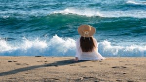 Rear view of an unrecognizable young woman with summer hat hat, sitting on the beach and watching waves and stormy sea. Nostalgic scene.