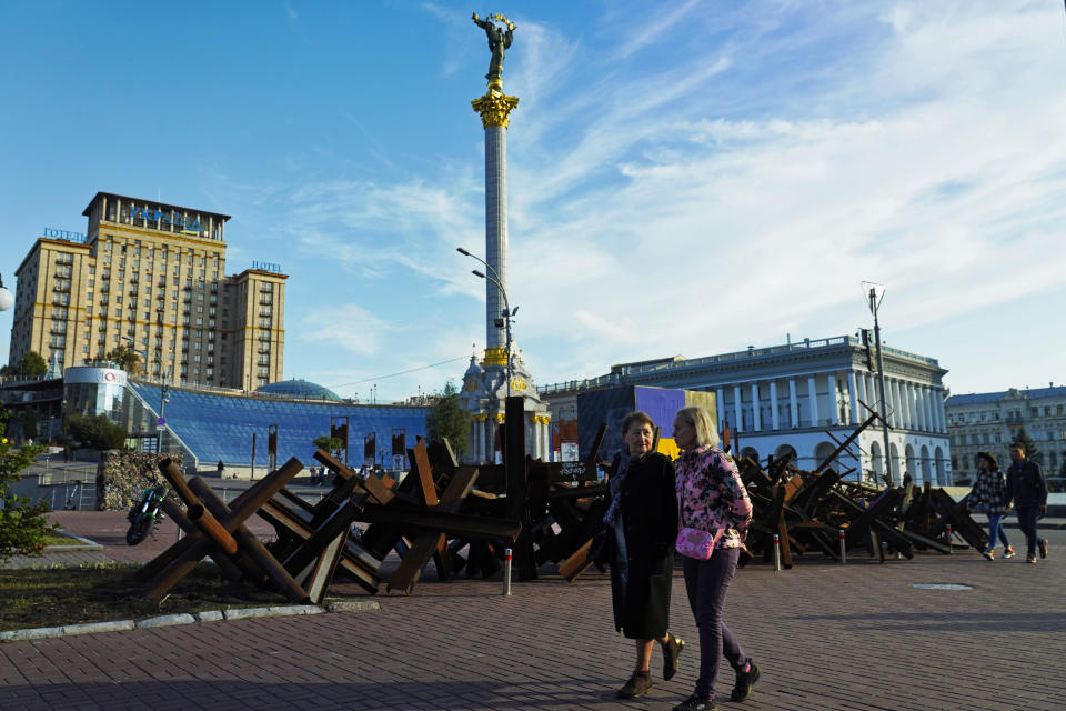 Two women walk by rusted hedgehogs once put in the streets to block tanks and the Independence Monument in Kyiv, Ukraine, Sunday, Sept. 11, 2022. (AP Photo/Jon Gambrell)