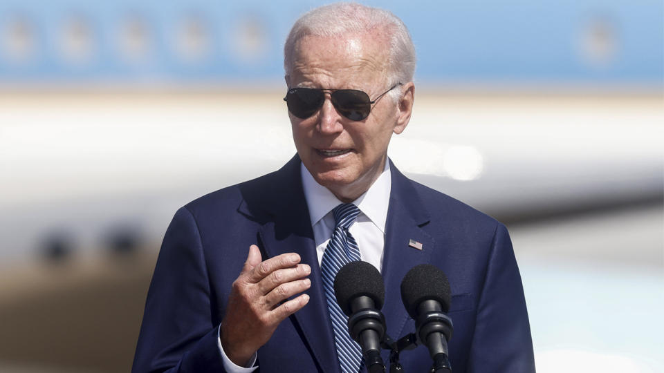 President Biden, wearing sunglasses, at a microphone on the tarmac with Air Force One in the background.