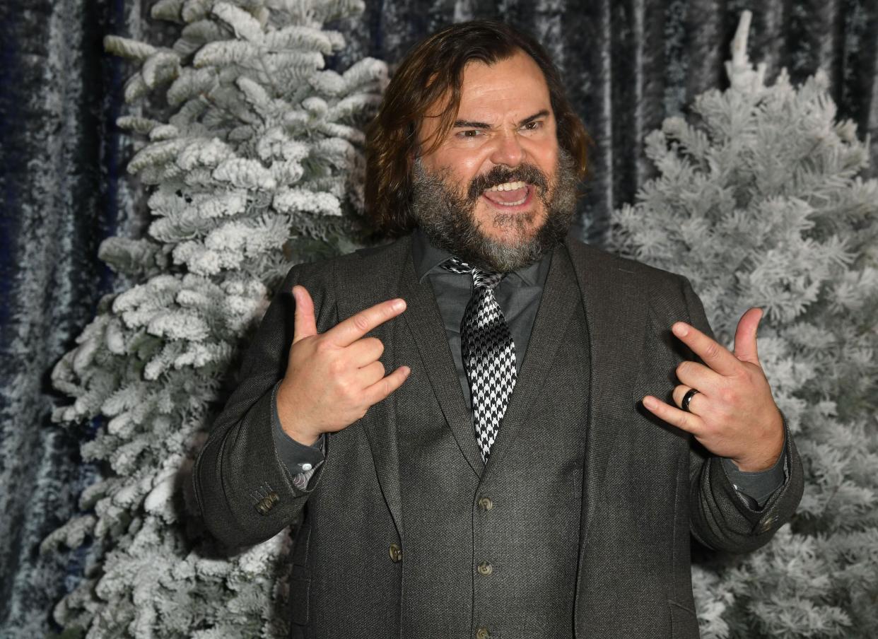 Jack Black doing the "Hook Em' Horns" finger signs in a stylish way. He is also sporting a well tailored grey suit with black and white poaches tie to match