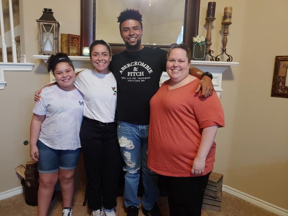 a family of four comprised of a youngest sister, middle sister, and older brother as well as their mother standing in front of a fireplace. the oldest brother has his arms around his sisters and mother