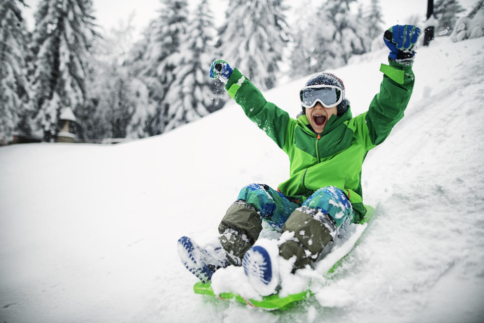 Enthusiastic child wearing ski goggles while sledding down a hill on a plastic sled with his arms in the air