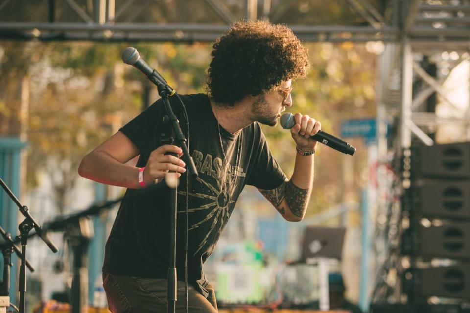 A man raps with a microphone held up to his face against a backdrop of trees.