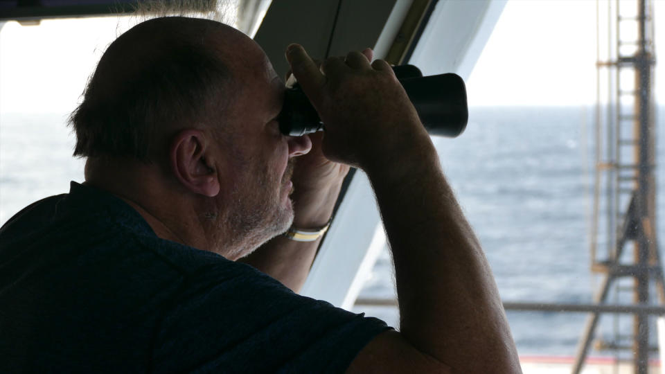 A man on a ship looks through binoculars.