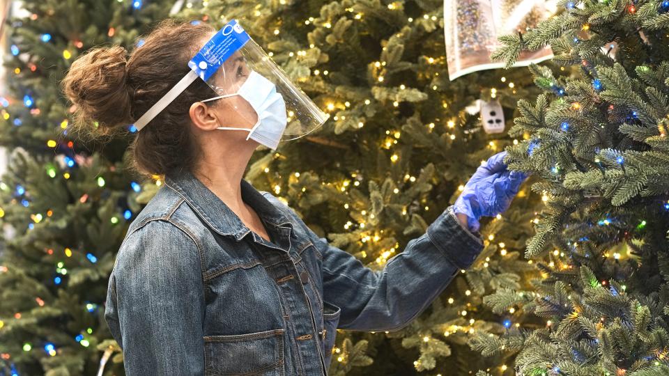 Mature caucasian woman shopping for Artificial Christmas Tree inside a megastore wearing a protective face mask, a face shield and protective gloves