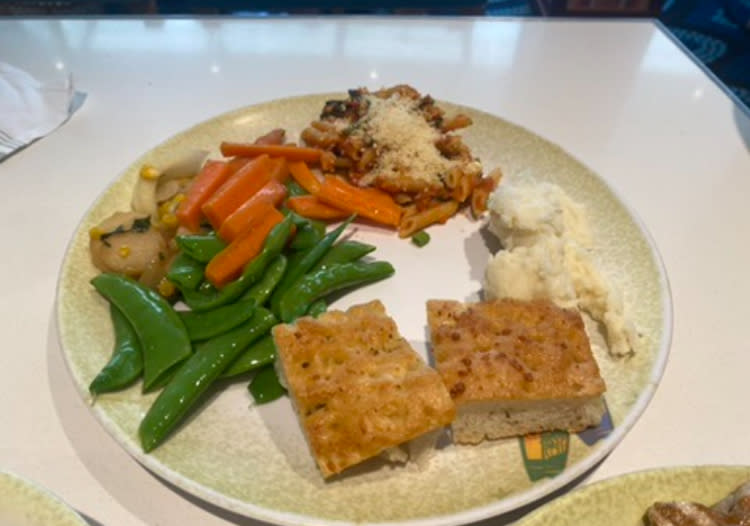 A dinner plate with sautéed vegetables, baked pasta topped with cheese, mashed potatoes, and two pieces of focaccia bread