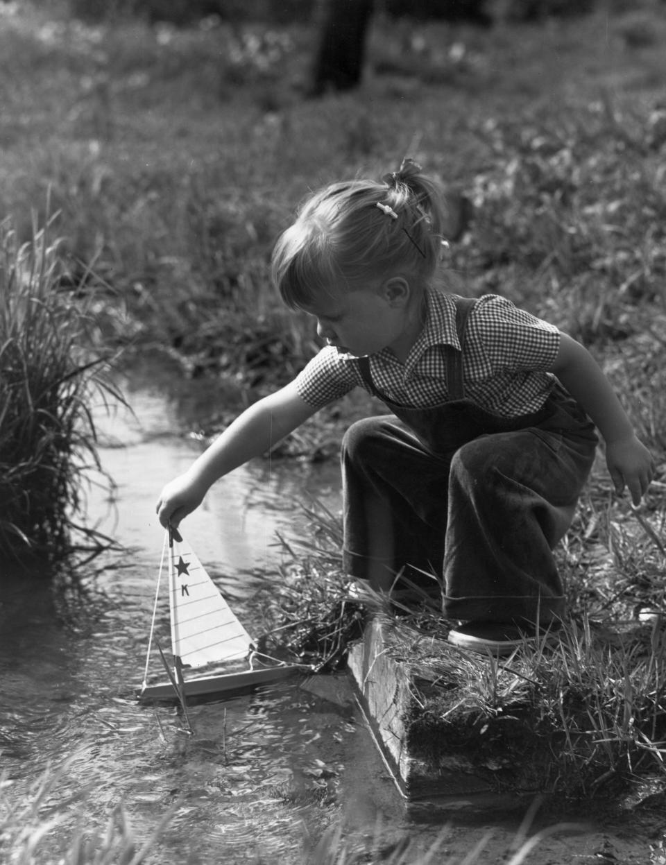 Circa 1945:  Full-length image of a young girl crouching on the grassy banks of a stream, holding the mast of a toy sailboat in the water.  (Photo by Lambert/Getty Images)