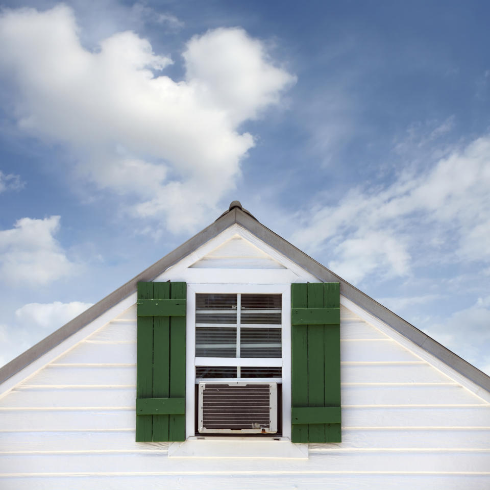 The image shows the top portion of a house with an air conditioner in a window and green shutters against a blue sky with clouds