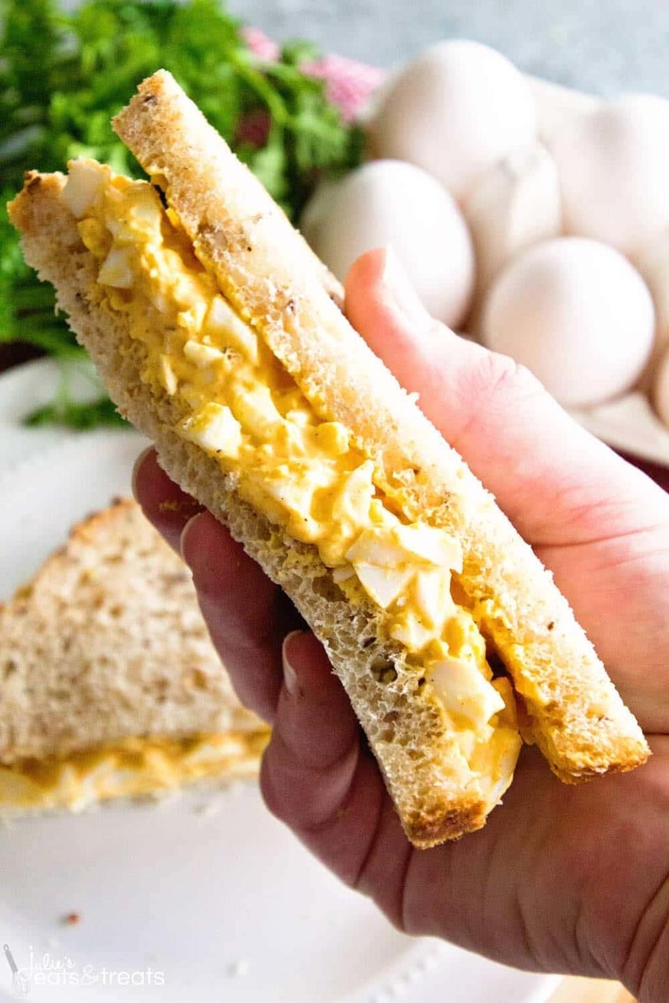 A hand holding a sandwich filled with egg salad. Other eggs and greenery are visible in the background