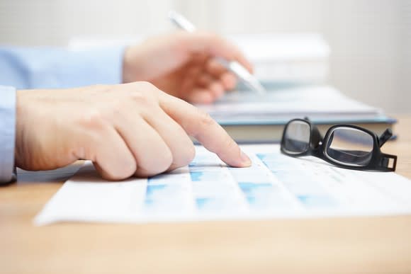 Man's hands pointing to a piece of paper with charts on it, which sits atop a desk. There's a pen in his hand and a pair of glasses on the charts.